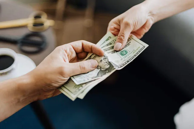 Două mâini țin și transmit un teanc de bancnote de dolar american deasupra unei suprafețe de lucru întunecate.