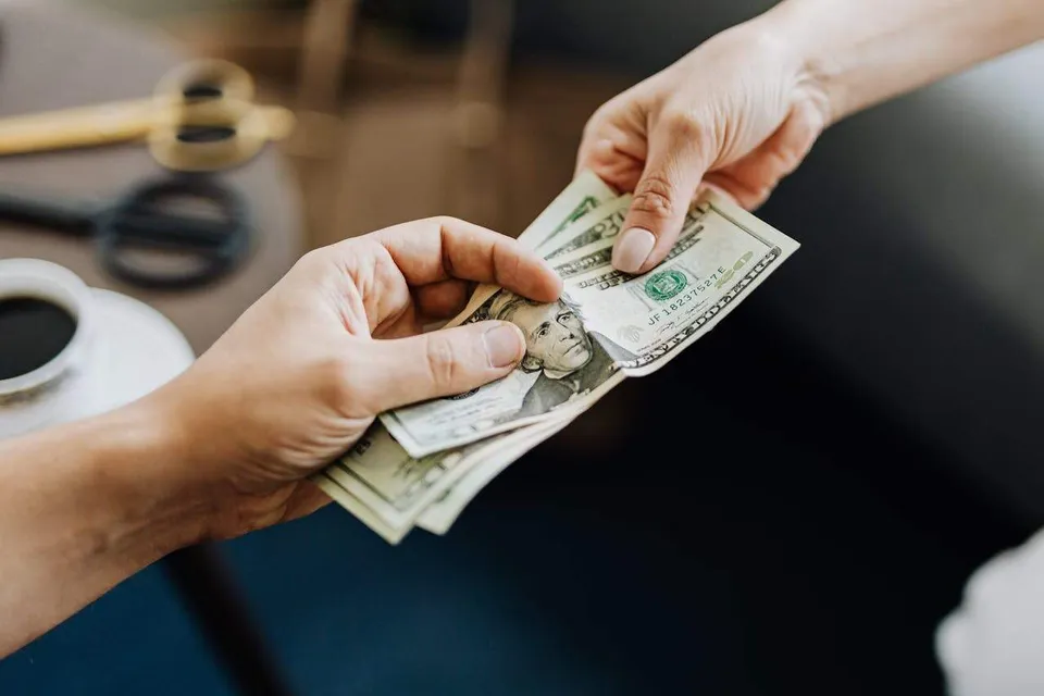 Două mâini țin și transmit un teanc de bancnote de dolar american deasupra unei suprafețe de lucru întunecate.