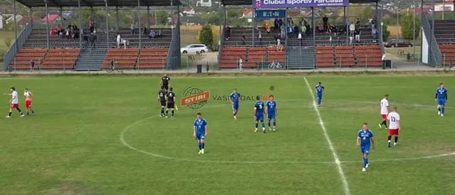 Jucători de fotbal în echipamente albastre și albe sunt pe un teren de fotbal verde, în fața unor tribune cu spectatori.
