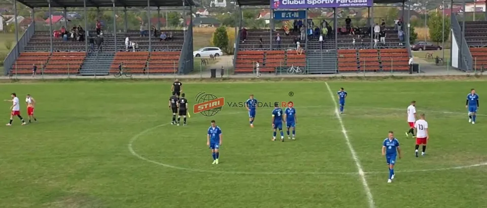 Jucători de fotbal în echipamente albastre și albe sunt pe un teren de fotbal verde, în fața unor tribune cu spectatori.