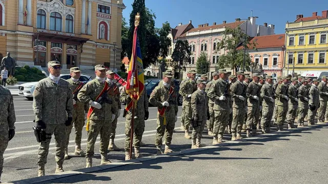 Soldați în uniforme de camuflaj stau în rânduri pe un drum asfaltat, în fața unor clădiri urbane, lângă un steag colorat.