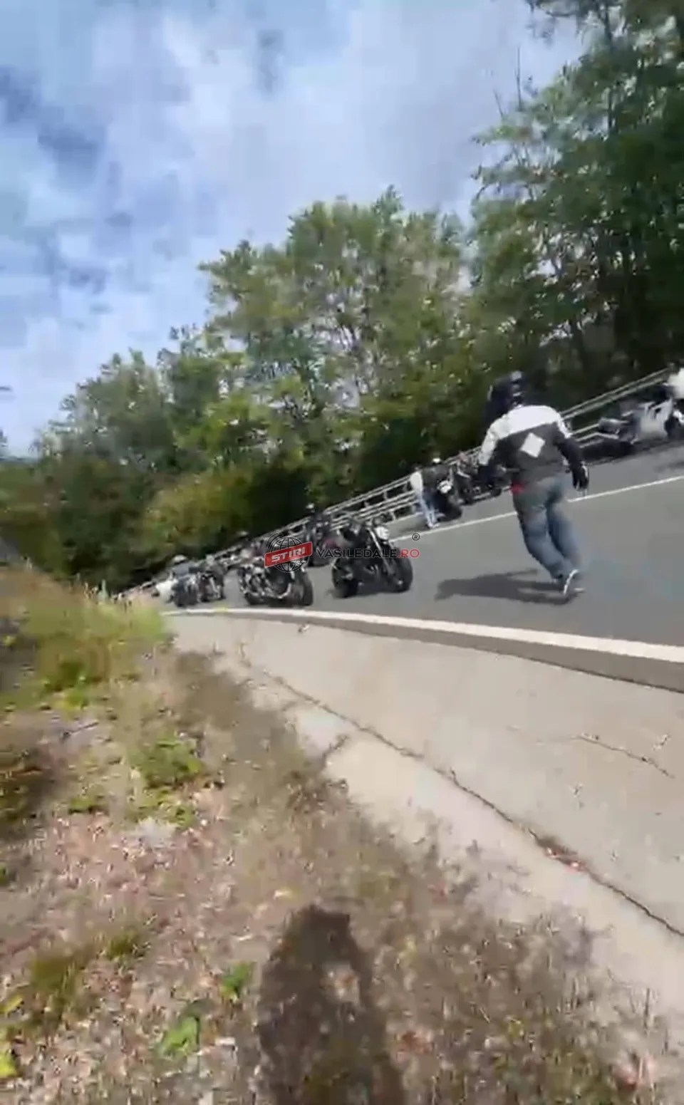 O persoană cu pantaloni albaștri și o jachetă albă merge pe un drum asfaltat lângă mai multe motociclete parcate.