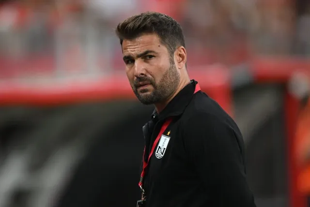 Un bărbat cu barbă și păr scurt, purtând un tricou negru cu un emblema pe piept, privește spre lateral într-un decor de stadion.
