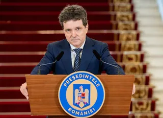 Un bărbat cu părul închis la culoare, purtând un costum albastru și cravată, stă în spatele unui podium de lemn cu un emblema circulară.