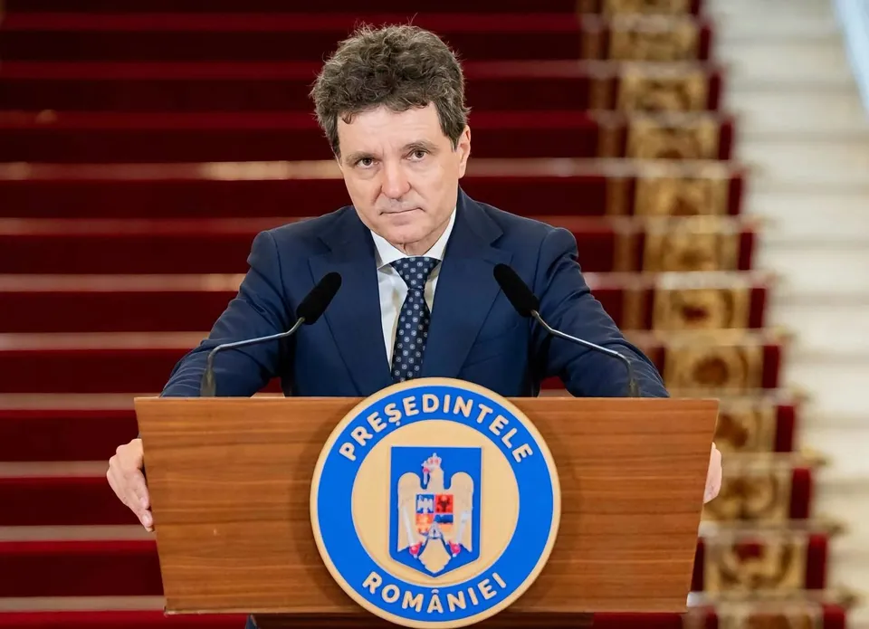 Un bărbat cu părul închis la culoare, îmbrăcat în costum albastru și cravată, stă în spatele unui podium de lemn cu un emblema circulară.