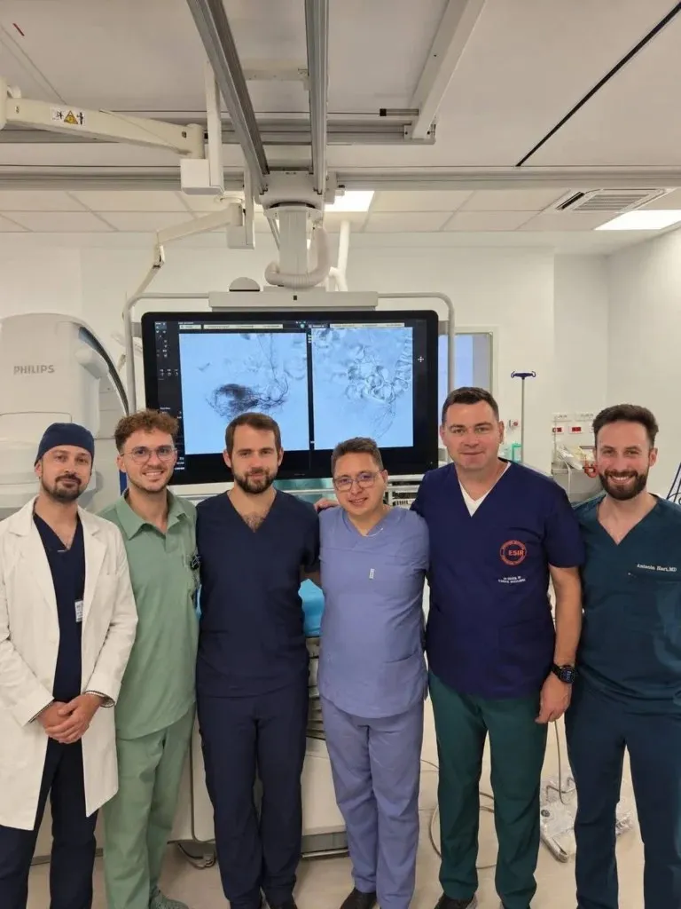 Șase persoane în halate medicale de diferite culori stau în rând într-o încăpere cu echipamente medicale și un monitor mare în fundal.