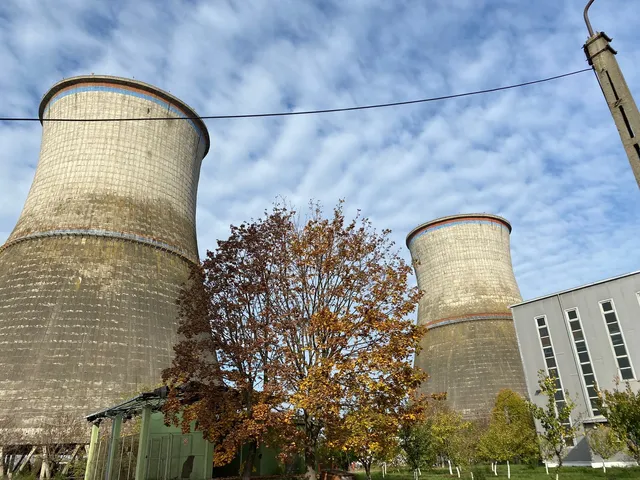 Două turnuri de răcire masive din beton sunt situate lângă un arbore cu frunze galbene și o clădire gri sub un cer înnorat.