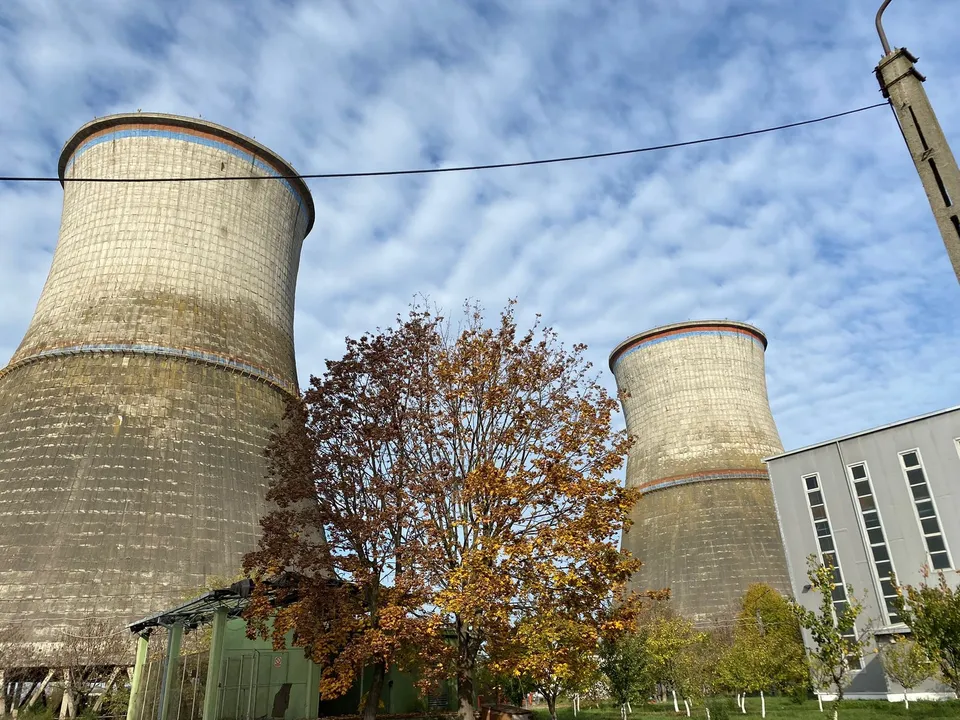 Două turnuri de răcire masive din beton sunt situate lângă un arbore cu frunze galbene și o clădire gri sub un cer înnorat.