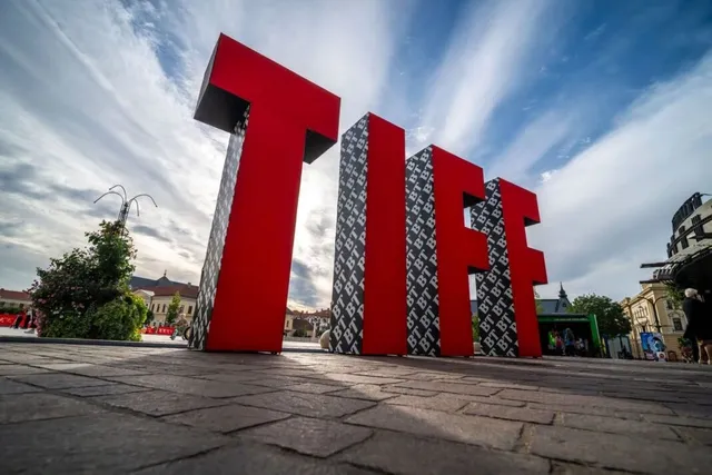 Literele mari, roșii și negre care formează textul TIFF sunt amplasate pe o suprafață pavată sub un cer cu nori subțiri.