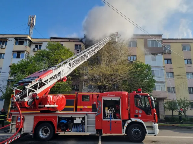 Un autospecial de pompieri roșu este parcat pe stradă, având o scară extensibilă ridicată spre un bloc de apartamente de lângă un copac.