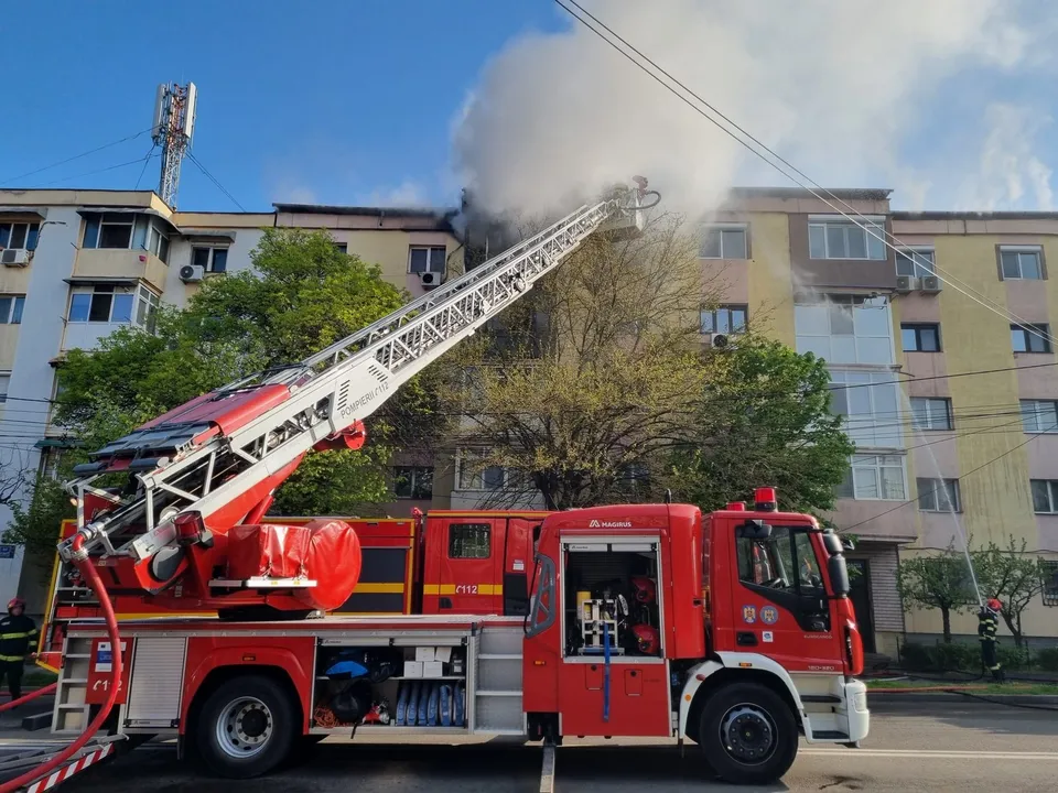 Un autospecial de pompieri roșu este parcat pe stradă, având o scară extensibilă ridicată spre un bloc de apartamente de lângă un copac.