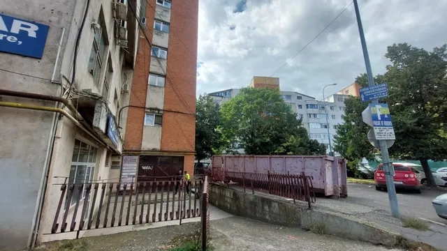 Un bloc de apartamente cu fațadă de cărămidă și ferestre albe stă lângă un gard metalic maro și un container roz.