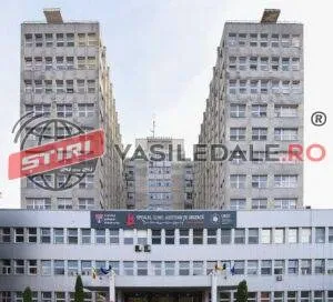 Două clădiri înalte de beton cu ferestre multiple sunt situate lângă o structură mai joasă sub un cer deschis.