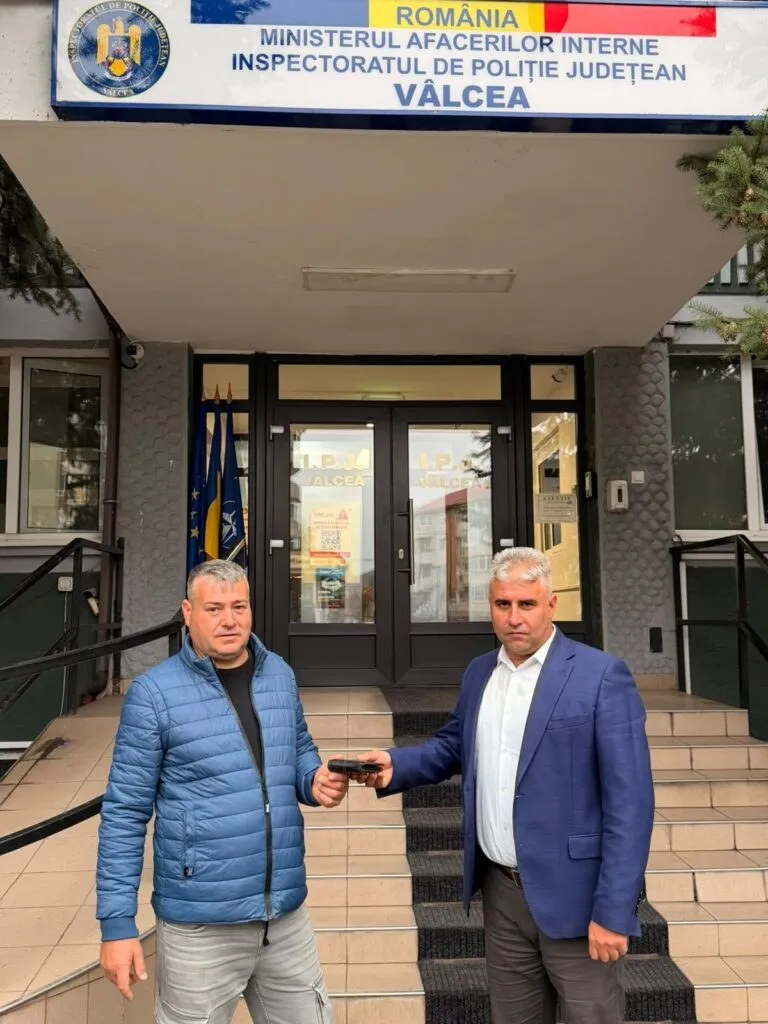 Doi bărbați stau pe scări în fața unei clădiri, unul purtând o jachetă albastră și celălalt un sacou albastru, ținând un obiect mic.