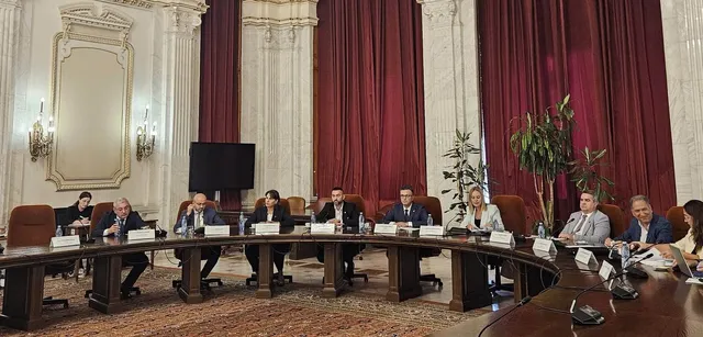 Oameni stând la o masă mare, curbată, într-o sală cu pereți albi, decorațiuni aurii și draperii roșii. Pe masă sunt sticle de apă și documente.