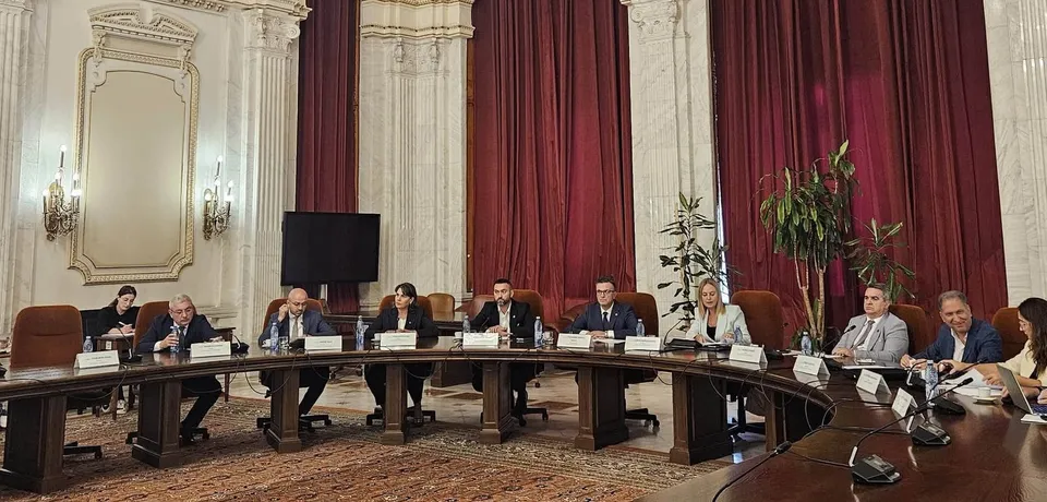 Oameni stând la o masă mare, curbată, într-o sală cu pereți albi, decorațiuni aurii și draperii roșii. Pe masă sunt sticle de apă și documente.
