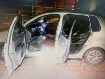 O mașină de culoare deschisă cu ambele portiere din partea pasajului deschise este parcată pe un pavaj lângă un zid de cărămidă.