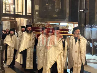 Cinci bărbați în haine liturgice albe și aurii poartă un obiect dreptunghiular decorat într-un interior cu coloane și ferestre înalte.