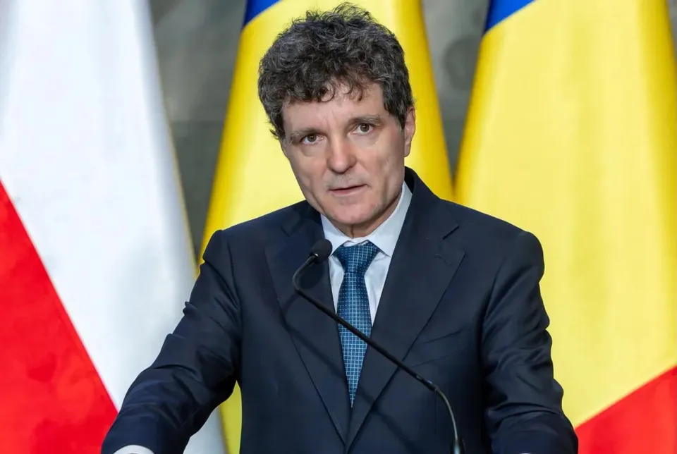 Un bărbat cu părul șaten și creț, purtând un costum închis la culoare, cămașă albă și cravată albastră, stă în fața unui microfon.