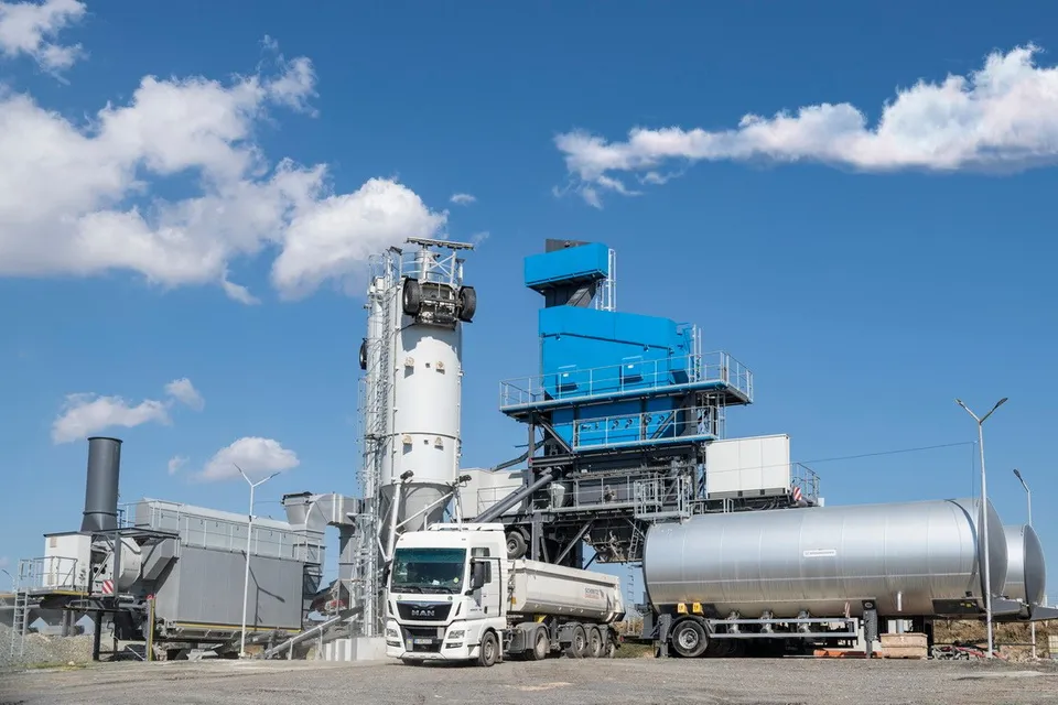 O stație industrială albastră și albă este situată lângă un camion alb și un vapor de transport gri sub un cer cu nori.