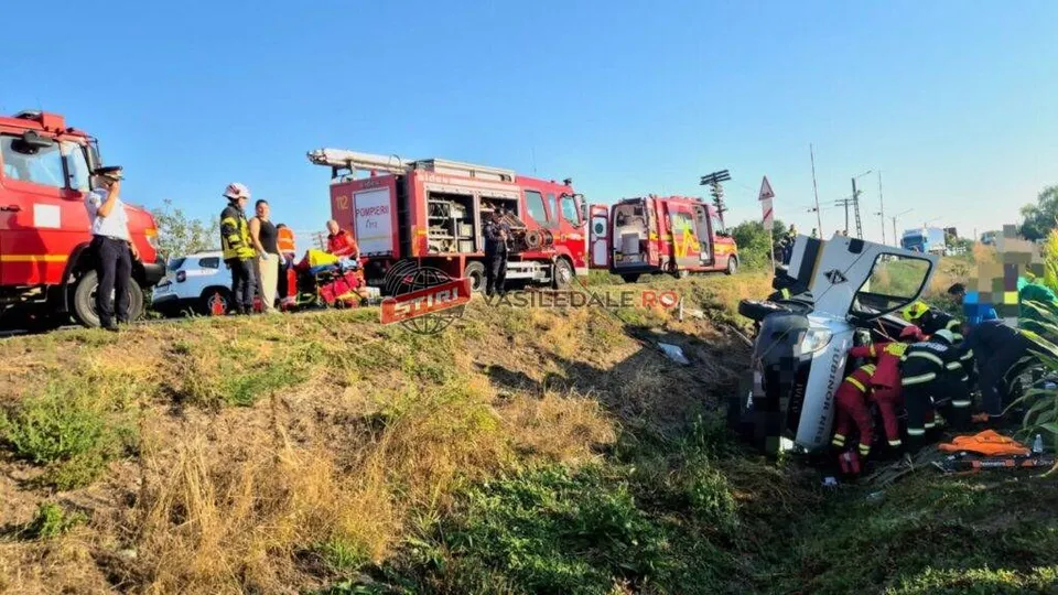 Echipaje de salvatori în uniforme colorate lucrează lângă un vehicul alb răsturnat într-un șanț, în prezența mai multor autospeciale de pompieri roșii.
