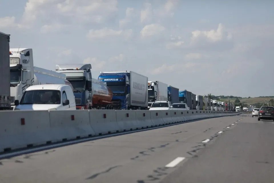 Mai multe camioane și autoutilitare sunt parcate pe o bandă de circulație lângă un parapet de beton sub un cer cu nori.