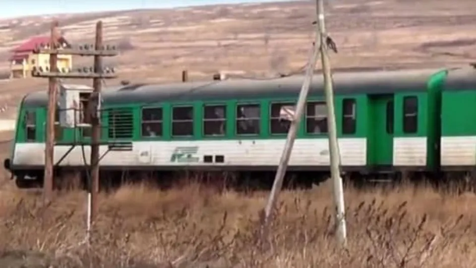 Un tren verde și alb circulă pe o cale ferată printre vegetație uscată și stâlpi de utilități.