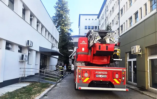 O autospecială de pompieri roșie, cu scara ridicată, este parcată lângă o clădire albă, cu mai mulți pompieri în uniformă lângă intrare.