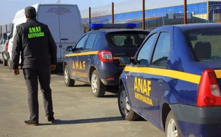 Un bărbat cu o vestă neagră pe care scrie ANAF ANTIFRAUDA stă lângă două mașini albastre marcate cu același text.
