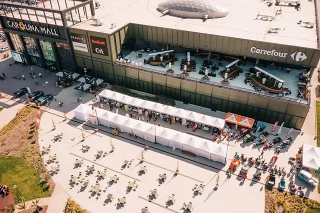 O vedere aeriană a unui centru comercial cu un parc auto și pavilioane albe dispuse pe o suprafață pavată în fața clădirii.