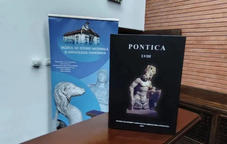 Un banner albastru cu imagini de clădiri și sculpturi stă lângă o carte neagră cu textul PONTICA LVIII pe o masă de lemn.