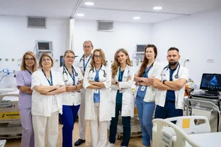 Opt persoane în halate albe și uniforme medicale stau în rând într-o încăpere de spital cu echipamente medicale în fundal.