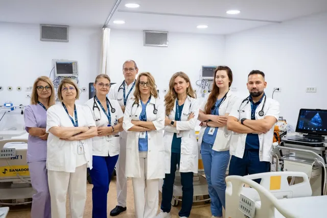 Opt persoane în halate albe și uniforme medicale stau în rând într-o încăpere de spital cu echipamente medicale în fundal.