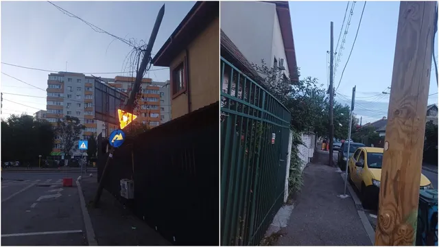 Un stâlp înclinat cu cabluri este sprijinit de un gard metalic lângă o clădire, în apropierea unui semn de circulație și al unui bloc.