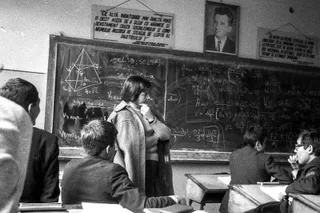 O sală de curs cu elevi așezați la bănci, o persoană în picioare lângă o tablă încărcată cu formule, desene geometrice și afișe suspendate.