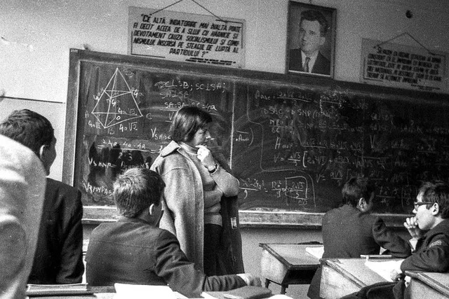 O sală de curs cu elevi așezați la bănci, o persoană în picioare lângă o tablă încărcată cu formule, desene geometrice și afișe suspendate.