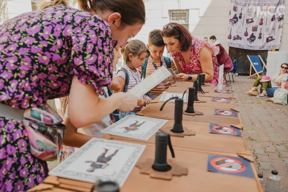 O femeie cu o bluză mov cu motive florale arată unor copii diverse obiecte de lemn și foi de hârtie așezate pe o masă lungă.