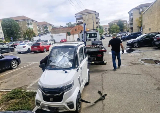 Un autovehicul mic de culoare albă este tras cu o remorcă de un camion de serviciu pe un drum asfaltat dintre clădiri de locuințe.