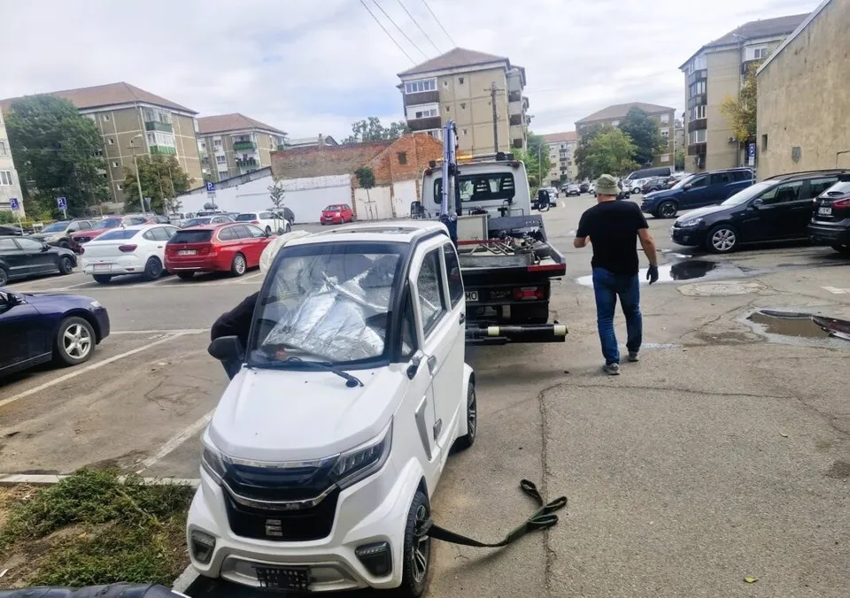 Un autovehicul mic de culoare albă este tras cu o remorcă de un camion de serviciu pe un drum asfaltat dintre clădiri de locuințe.