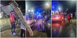 O topor cu mâner de lemn este ținut în prim-plan, în timp ce în fundal se văd persoane și vehicule de serviciu noaptea.
