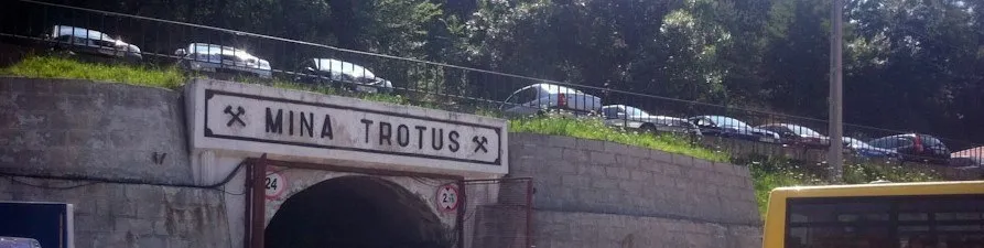 Un semn cu textul MINA TROTUS este montat deasupra unei intrare de tunel, sub un versant de deal cu mașini parcate pe marginea drumului.