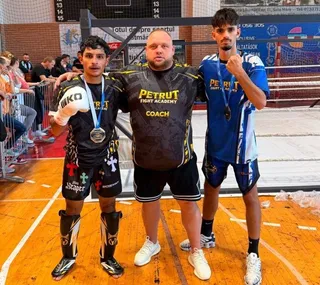 Trei bărbați stau în picioare pe un teren de sport, doi purtând medalii și tricouri cu inscripții, iar unul purtând un tricou de antrenor.