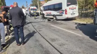 Un microbuz alb cu inscripții roșii și negre stă pe un drum lângă un vehicul de serviciu și persoane în uniformă.