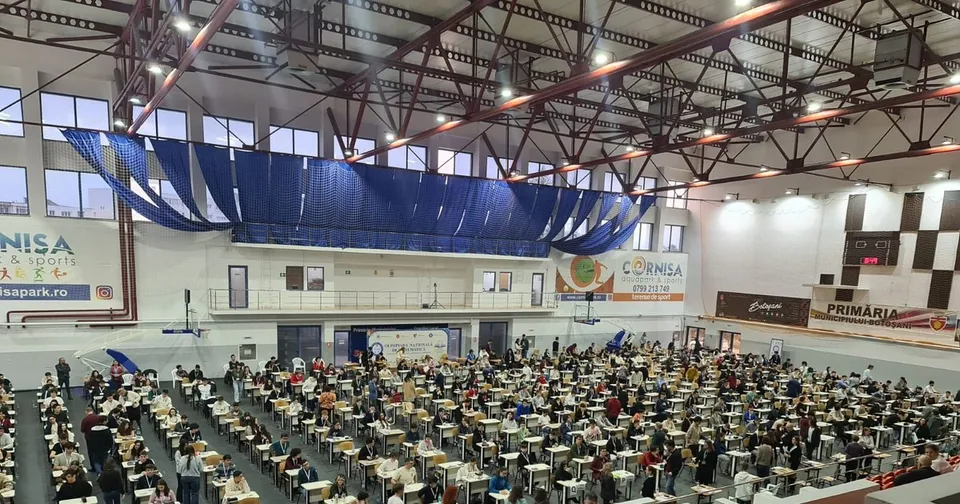 O mulțime de persoane stau la masa de lucru în rânduri organizate într-o sală de sport mare cu structură metalică și draperii albastre suspendate.