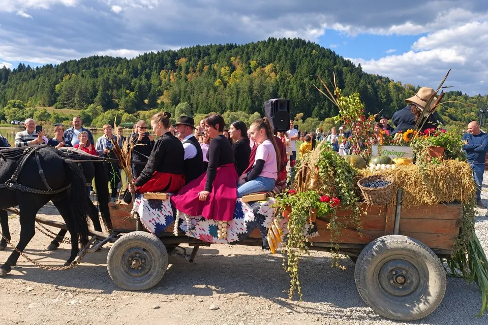 Un căruț decorat cu flori și plante este tras de un cal, având persoane așezate pe el în fața unui deal împădurit.
