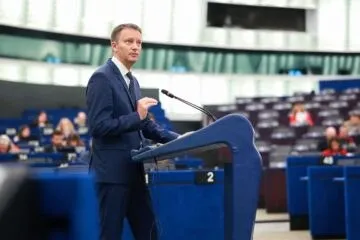 Un bărbat într-un costum albastru stă în spatele unui pupitru într-o sală cu multe scaune dispuse în rânduri.
