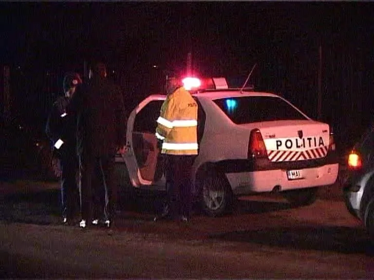 O mașină albă cu inscripția POLITIA este parcată pe un drum noaptea, lângă un vehicul cu portierele deschise și trei persoane în apropiere.