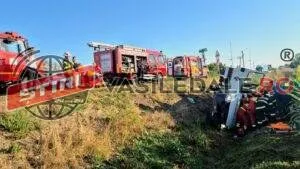 Mai multe autospeciale de pompieri roșii sunt parcate pe un teren cu iarbă uscată lângă un vehicul accidentat.