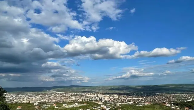 Un cer albastru cu nori albi și cenușii deasupra unui peisaj urban întins și unor dealuri verzi în fundal.
