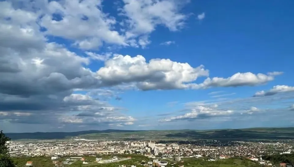 Un cer albastru cu nori albi și cenușii deasupra unui peisaj urban întins și unor dealuri verzi în fundal.